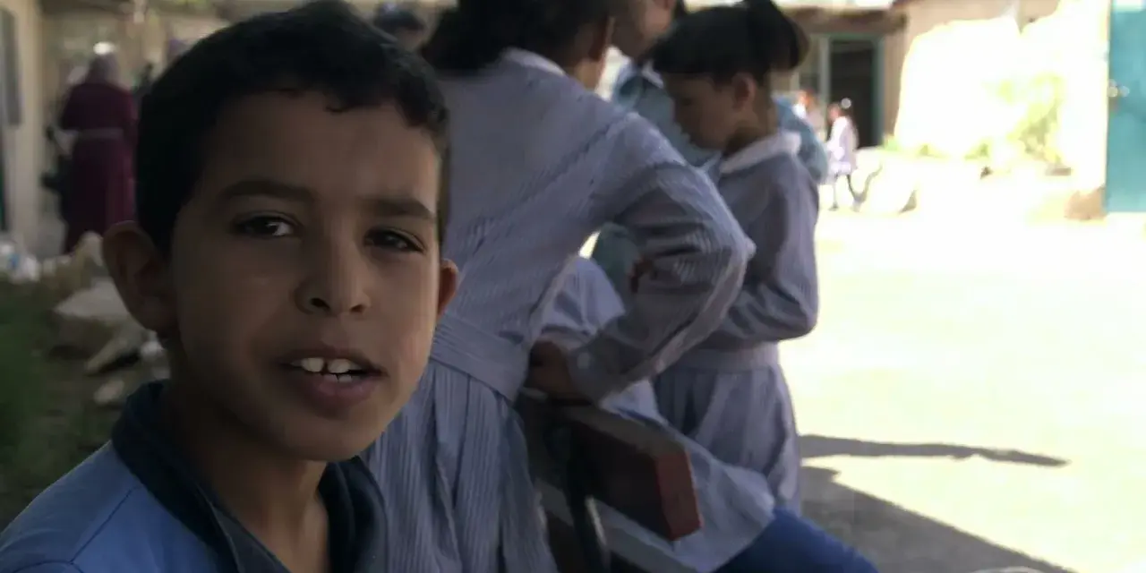 Un garçon souriant pose devant des filles en uniforme dans la même cour d'école.