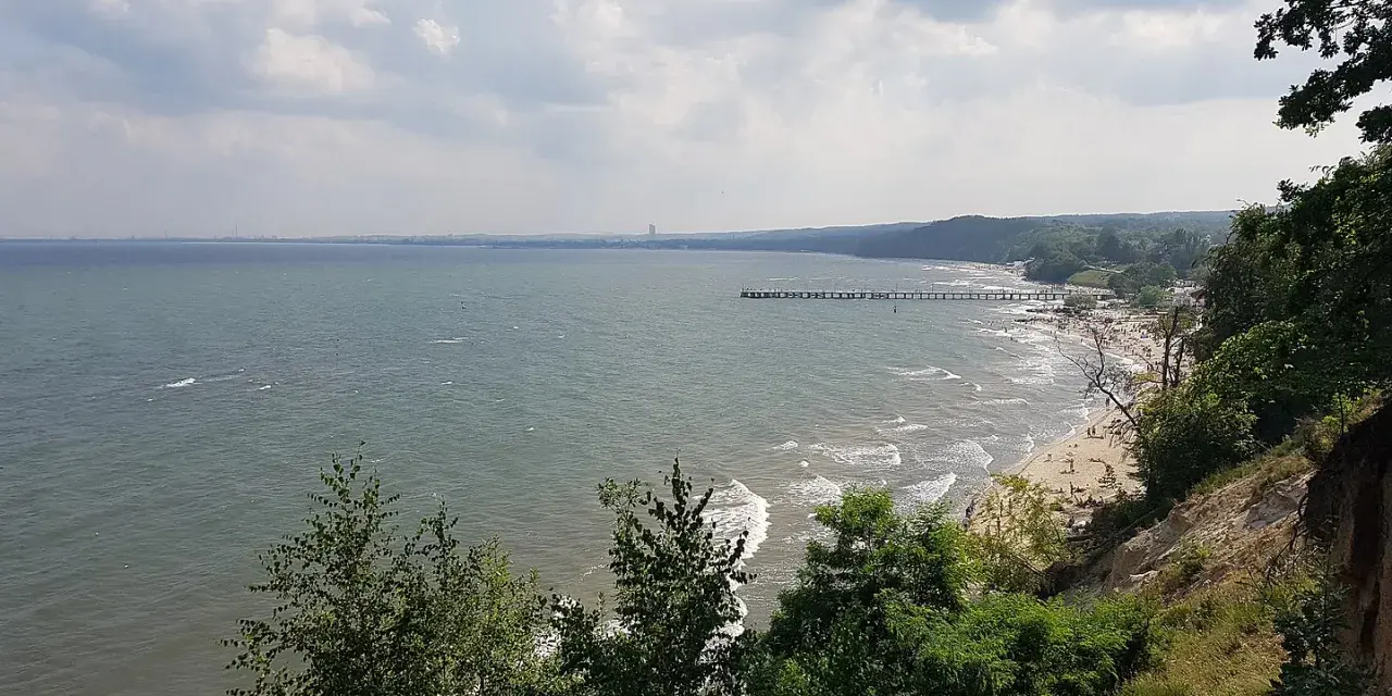 Widok z klifu orłowskiego na morze, plażę z ludźmi i molo. W oddali widać zarys wybrzeża.
