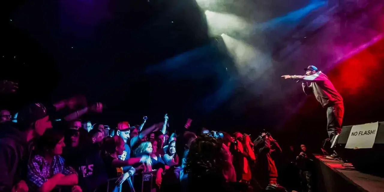 Raper na scenie, w świetle reflektorów, wskazuje palcem na tłum. Koncert w **3 wymiar**ze, pełen energii i emocji.
