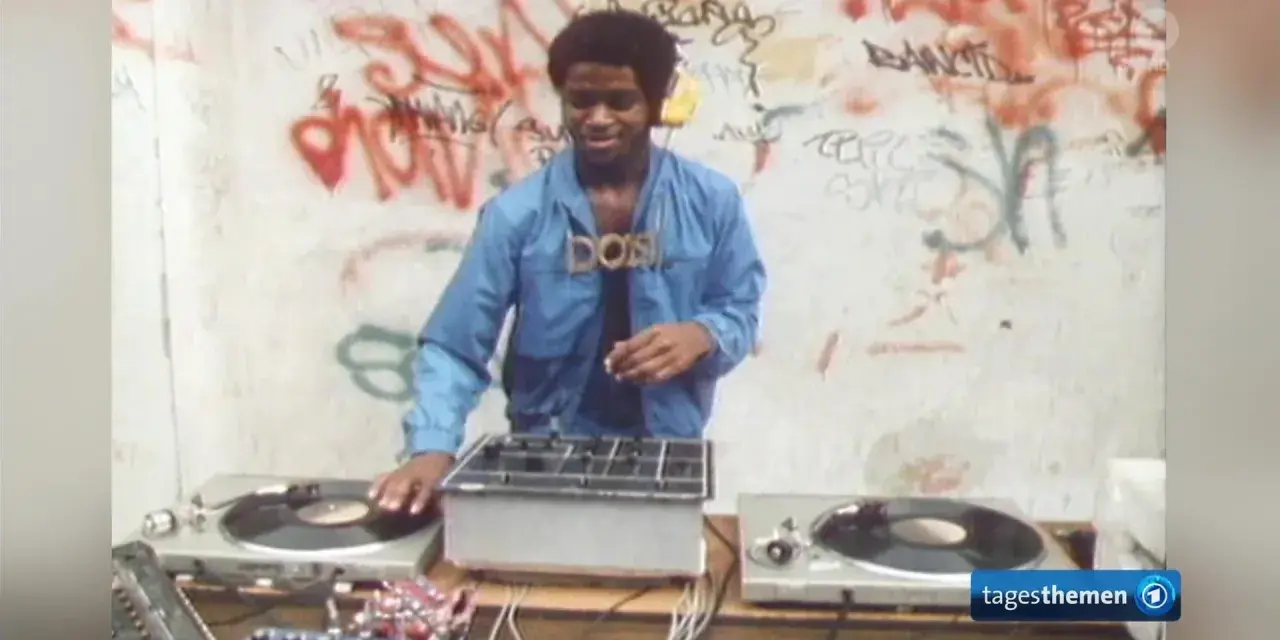 DJ Kool Herc, der oft als derjenige gilt, der Hip Hop erfunden hat, legt auf. Graffiti zieren die Wand im Hintergrund.