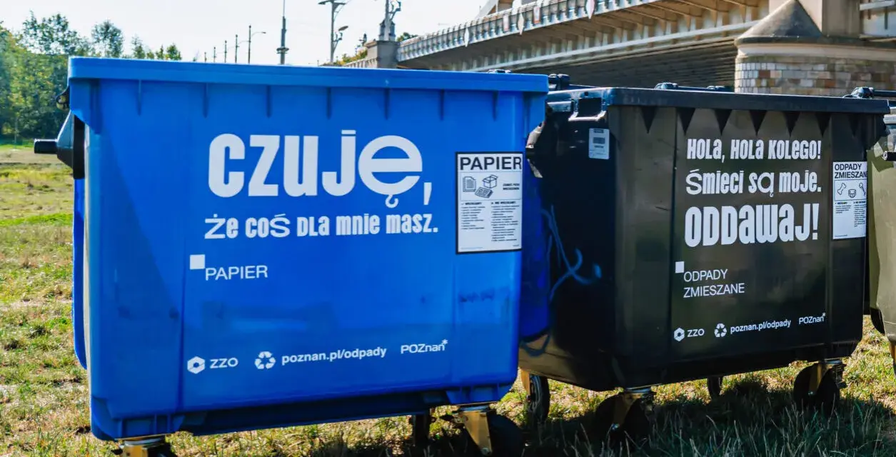 Niebieski kontener na makulaturę z napisem "czuję, że coś dla mnie masz. PAPIER" obok czarnego na odpady zmieszane.