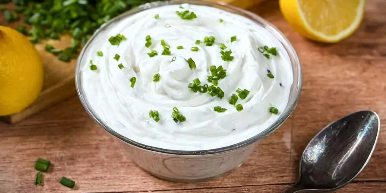 Cremige Sour Cream, garniert mit Schnittlauch, neben Zitronenspalten und einem Löffel auf Holzuntergrund.