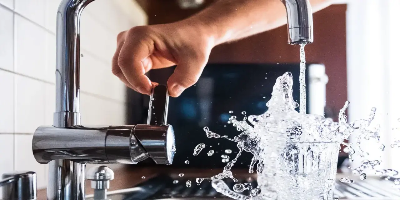 Mano girando la llave para cuidar el agua, que salpica al llenar un vaso.