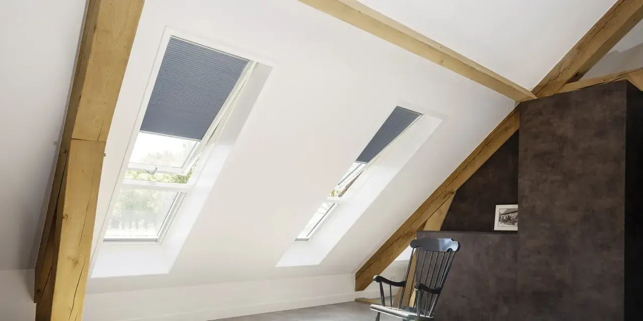 Schicke Dachfenster mit Plissees verkleiden. Holzbalken und ein gemütlicher Schaukelstuhl schaffen eine warme Atmosphäre.