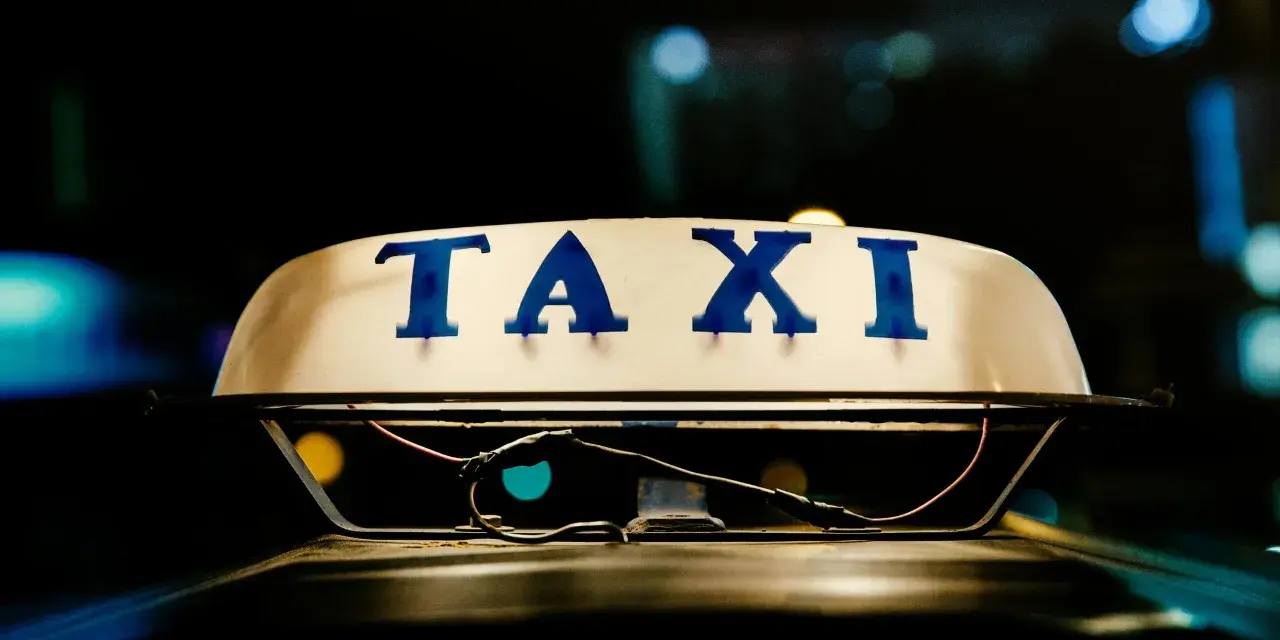 Tetto di un taxi con la scritta "TAXI" in blu. La luce soffusa e i riflessi suggeriscono la notte, un momento in cui ci si interroga quanto guadagnano i tassisti.