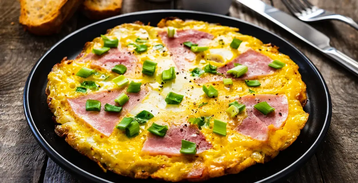 Pyszny omlet z szynką i serem, posypany szczypiorkiem, podany z grzankami. Idealny na śniadanie.
