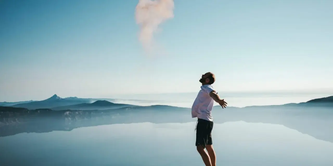 Mann mit ausgebreiteten Armen auf einem Felsvorsprung, der Rauch in den Himmel stößt. Ein Moment der Freiheit, wie Gottes Gunst.