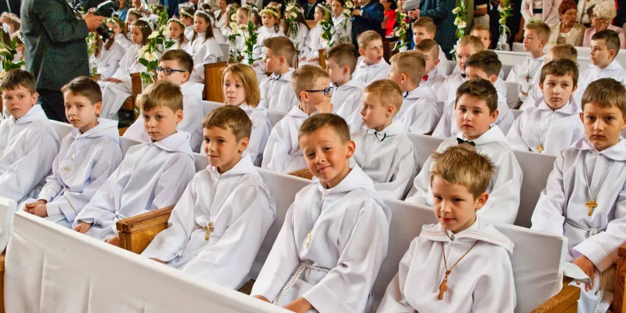 Dzieci w bieli, gotowe na rocznicę komunii świętej, siedzą w kościele. W tle widać zgromadzonych gości i fotografa.