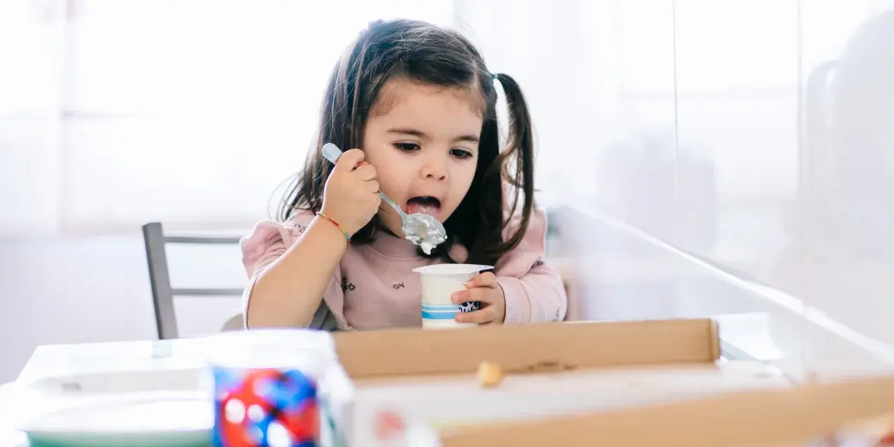 Mała dziewczynka z kitkami je jogurt z kubeczka. Od kiedy dziecko może jeść danonki? Odpowiedź jest prosta: gdy tylko zaczyna jeść pokarmy stałe.