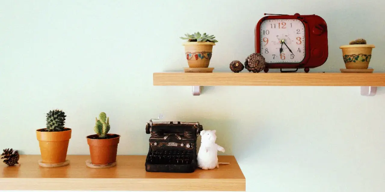 Ideas para decorar baldas salón: dos estantes de madera con cactus, una máquina de escribir antigua, un peluche blanco y un reloj rojo.