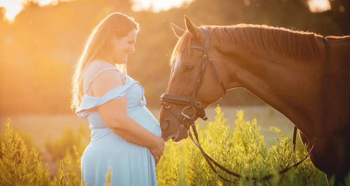 Une femme enceinte en robe bleue caresse un cheval brun au coucher du soleil. L'équitation enceinte est une belle façon de célébrer la maternité.