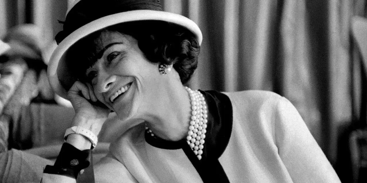 Coco Chanel sorride con un cappello e perle, un'icona di stile che sembra raccontare la storia di quando è nata Coco Chanel.