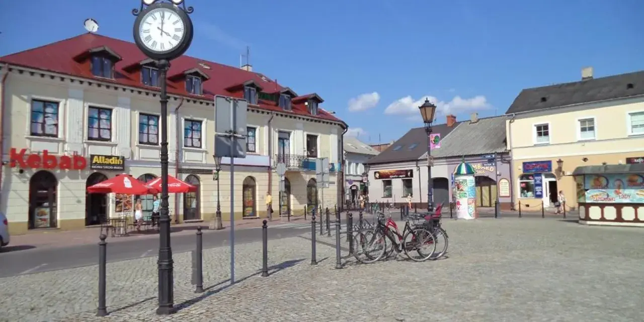 Słoneczny stary rynek łowicz z brukowanym placem, budynkami z czerwonymi dachami i zegarem na słupie.