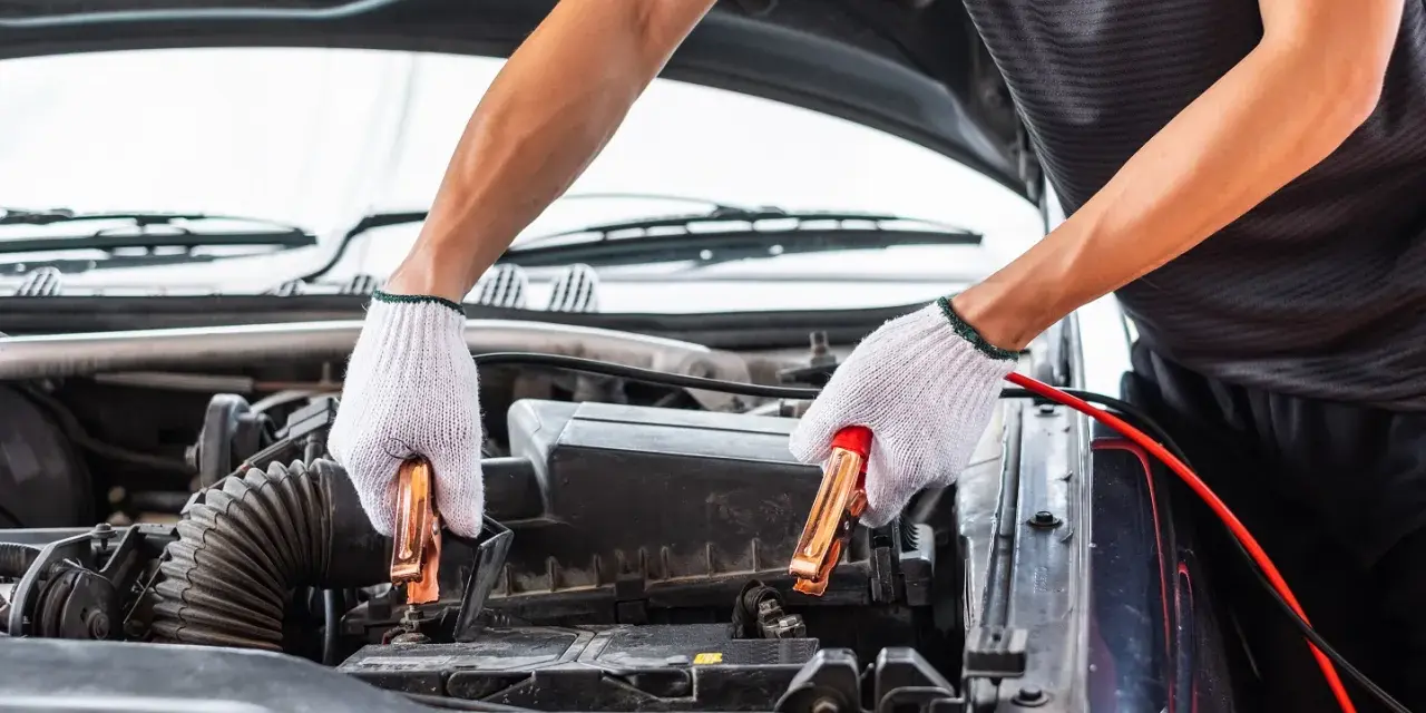 Hands in white gloves connect jumper cables to a car battery. This process helps answer how long does it take to charge a car battery.