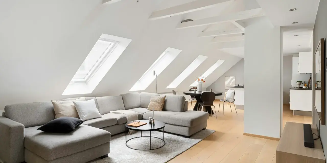Soggiorno mansardato con divano angolare grigio, tavolino nero e travi a vista moderne dipinte di bianco che creano un'atmosfera luminosa.