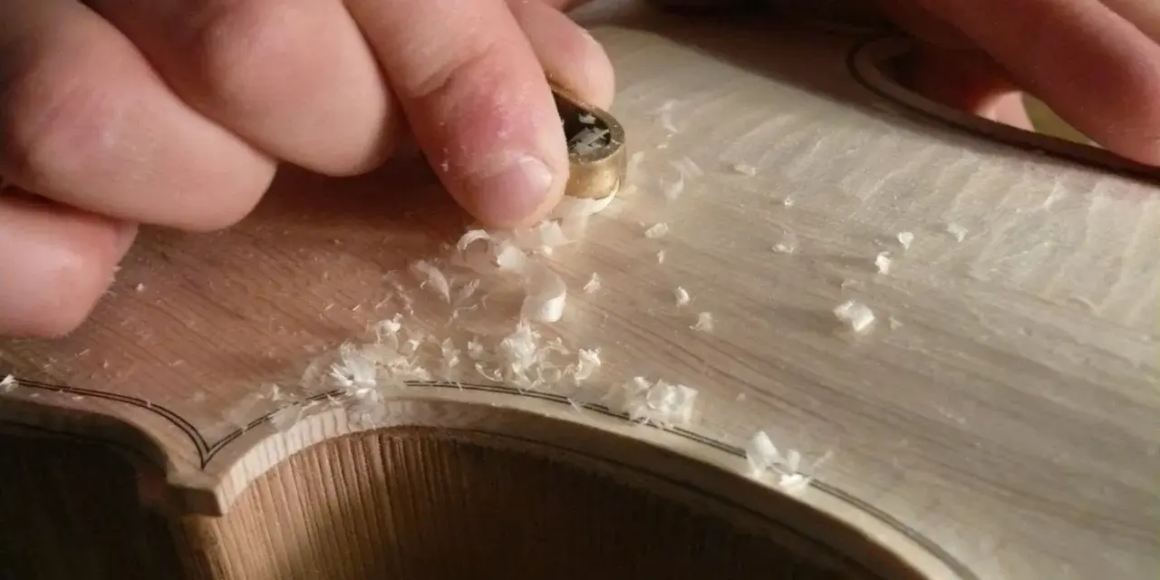 Hände bearbeiten mit einem Werkzeug das helle Holz einer Geige, Späne fliegen. Die Frage "aus welchem Holz besteht die Geige" wird hier visuell beantwortet.