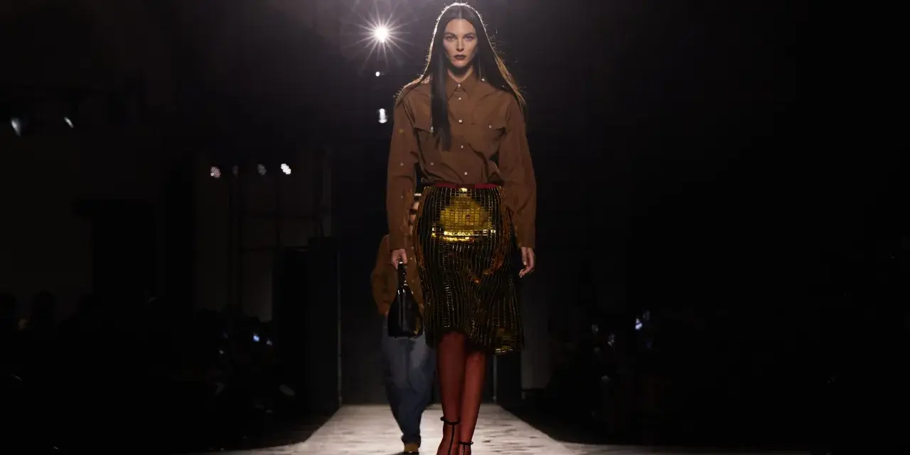 Modella su passerella, con gonna dorata e camicia marrone, un omaggio all'eleganza senza tempo di Prada, fondatore di un impero della moda.