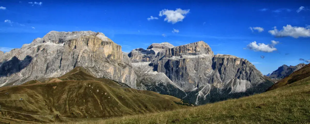 Vaste cime rocciose del gruppo Sella Dolomiti sotto un cielo azzurro con nuvole bianche. Prati verdi e sentieri serpeggiano sui pendii.