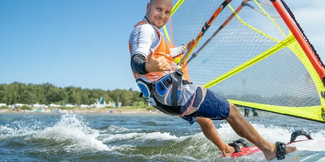 Mężczyzna na desce windsurfingowej, uśmiecha się i pokazuje kciuk w górę.