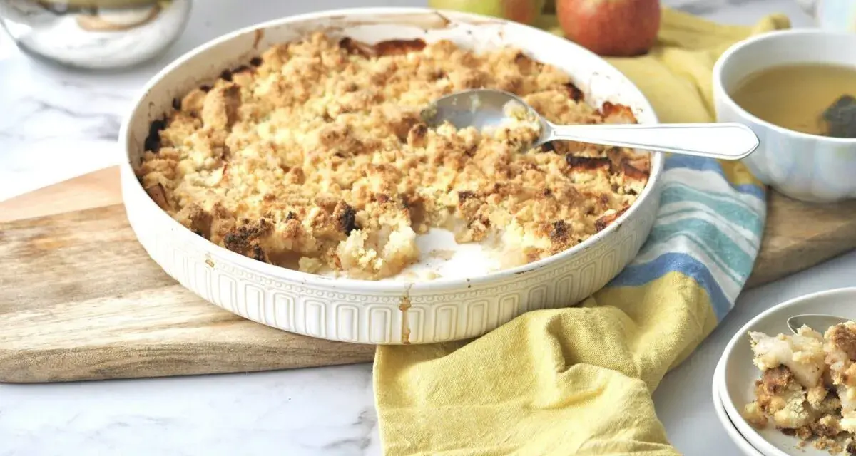 Ein goldbrauner Apfel-Crumble in einer Auflaufform, bereit zum Servieren. Daneben eine Tasse Tee und frische Äpfel.