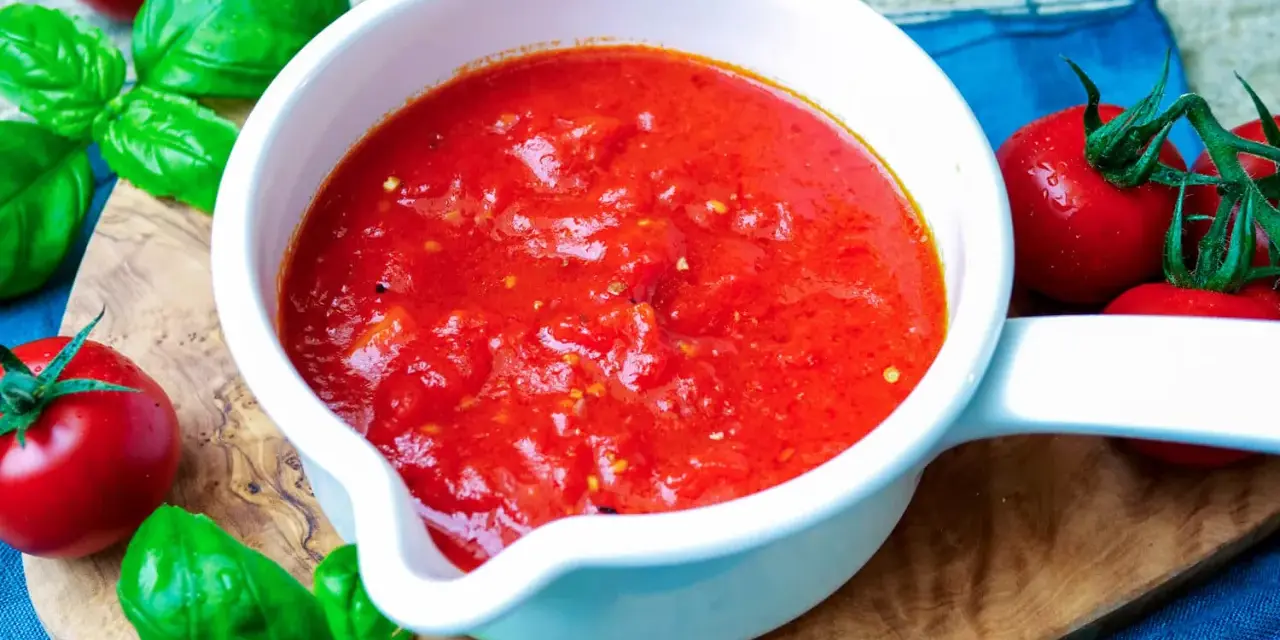 Frische Tomatensauce selber machen: Ein weißer Topf mit leuchtend roter Sauce, umgeben von reifen Tomaten und Basilikum auf einem Holzbrett.