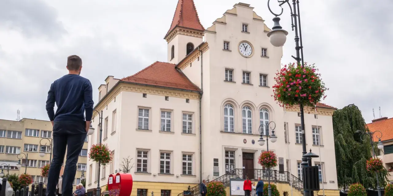 Mężczyzna podziwia ratusz, jedną z głównych trzebnickich atrakcji. W tle kwitnące pelargonie i zabytkowa architektura.