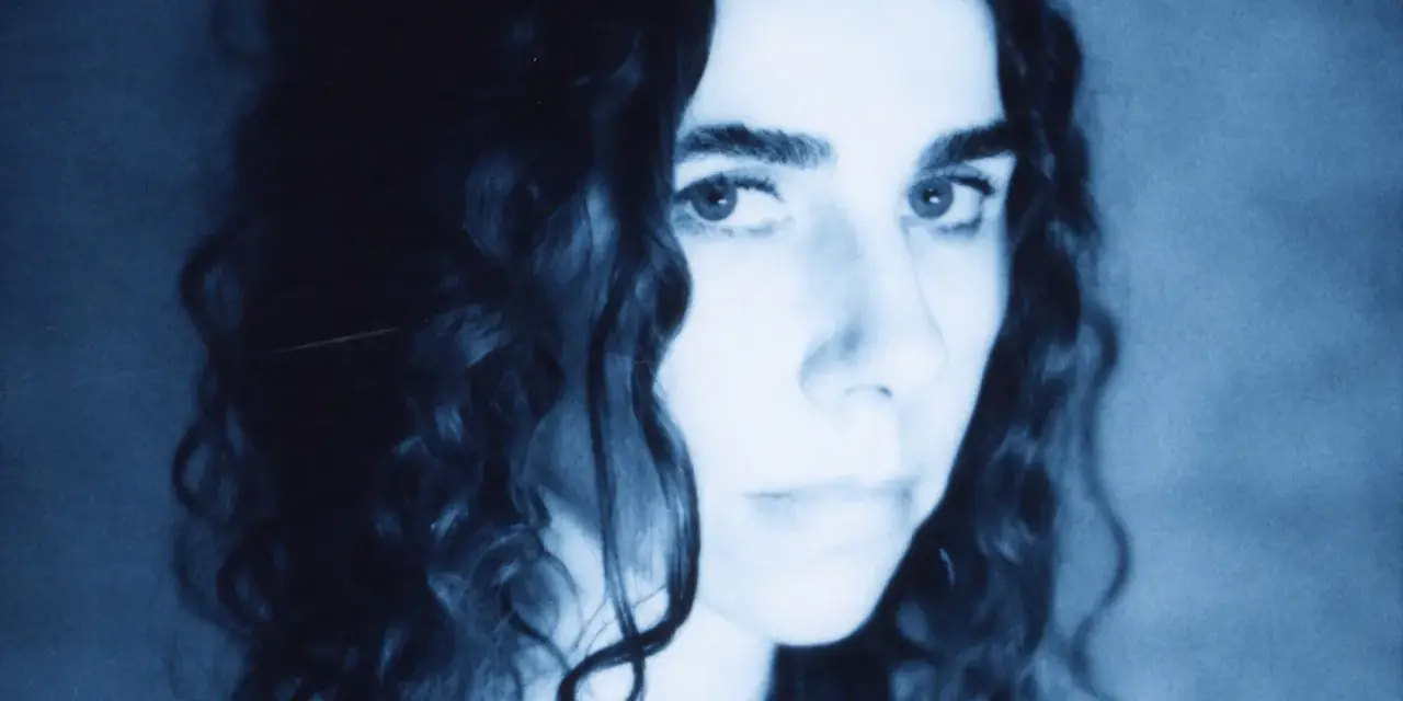 PJ Harvey, z kręconymi włosami, patrzy intensywnie. Jej twarz, w odcieniach niebieskiego, zdaje się opowiadać historię z "I Inside the Old I Dying".
