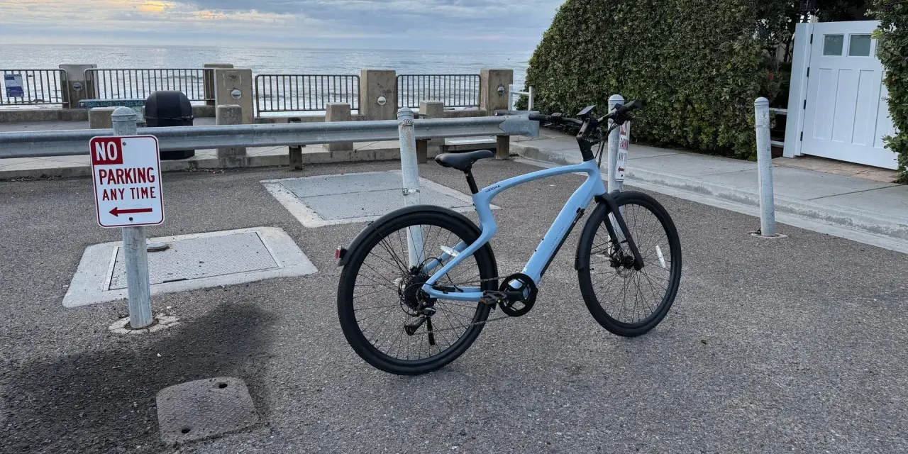 A light blue electric city bike is parked on asphalt near a "No Parking" sign, with the ocean and cloudy sky in the background.
