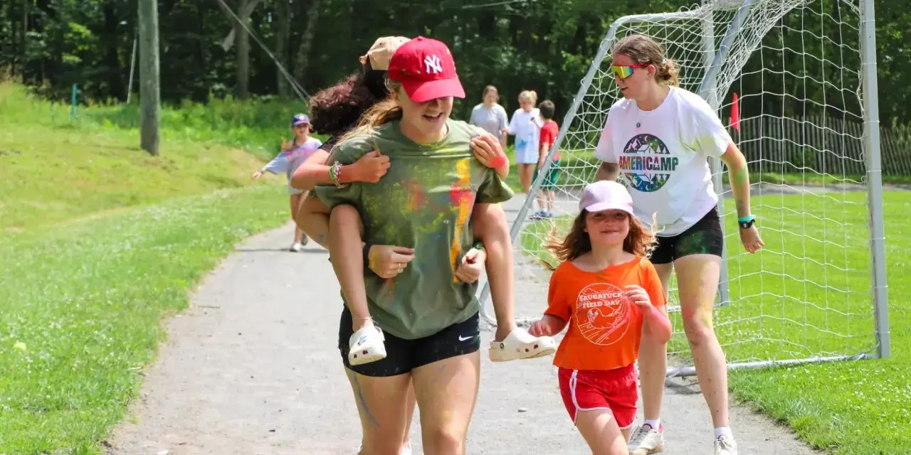 Mädchen haben Spaß bei einem Sommercamp in den USA. Eine trägt die andere auf dem Rücken, während sie lachend über einen Weg laufen.