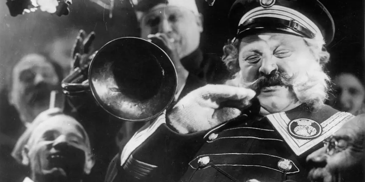 Un uomo con baffi e barba folta suona una tromba, circondato da volti sorridenti. Un'atmosfera da kammerspiel, un dramma intimo e intenso.