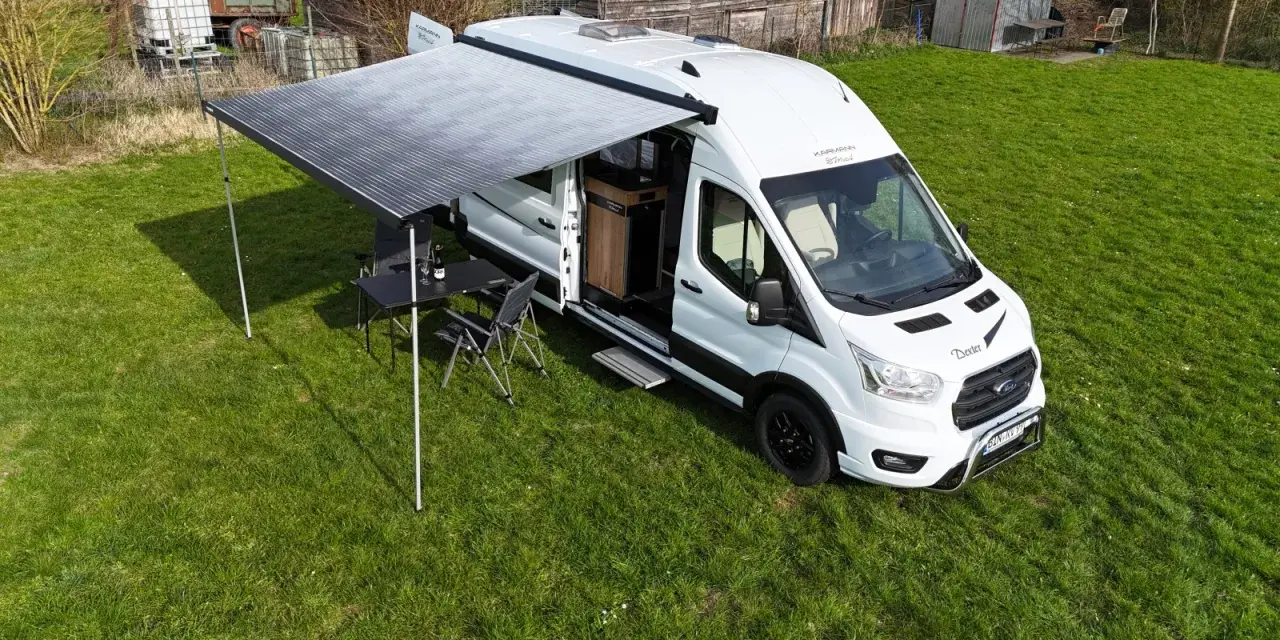 Vit husbil, Karmann Dexter, med utdraget markis och campingmöbler på gräs.