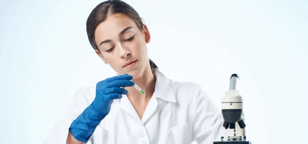 Giovane tecnologo alimentare in camice bianco, con guanti blu, analizza un campione con un contagocce accanto a un microscopio.