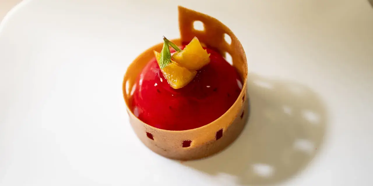 A red berry mousse dessert with orange zest and mint, presented in a decorative pastry shell on a white plate.