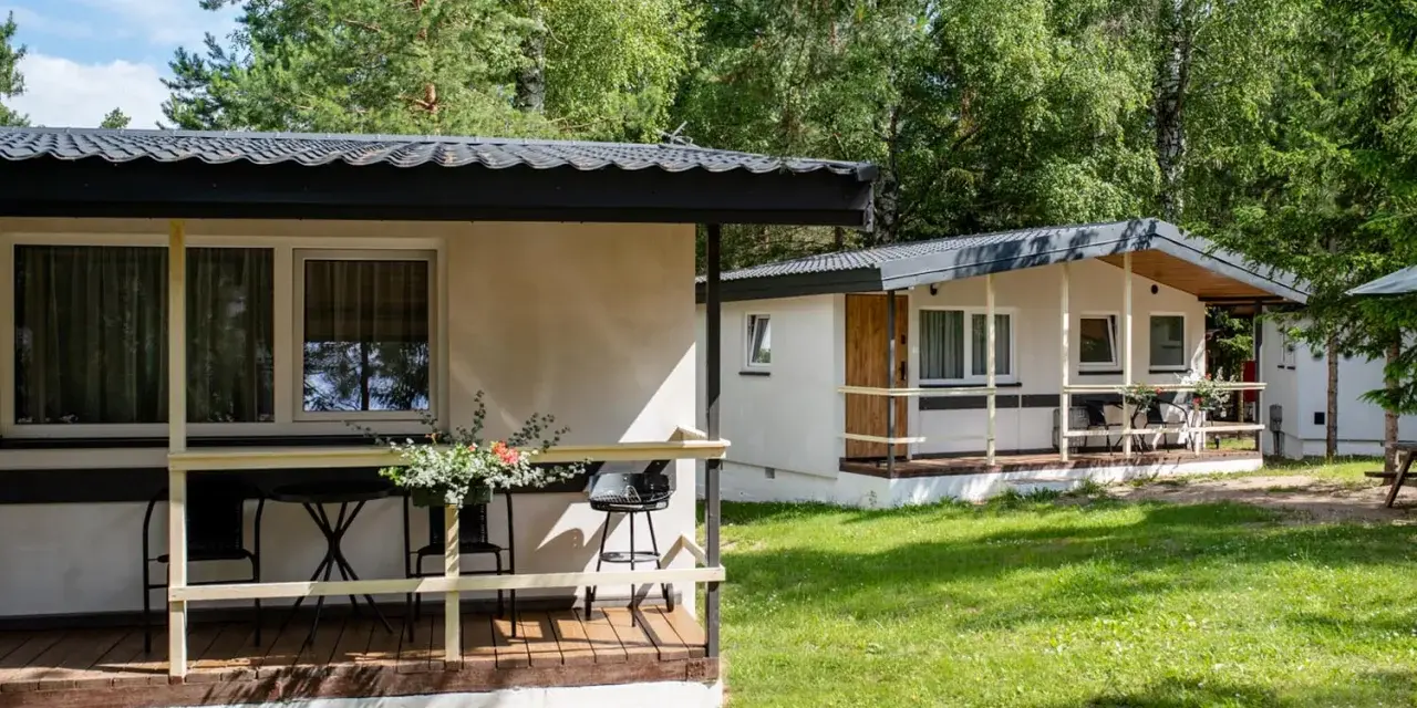 Dwa białe domki kempingowe w lesie, z werandami i meblami ogrodowymi. Idealne miejsce dla każdego **camping wagabunda**.