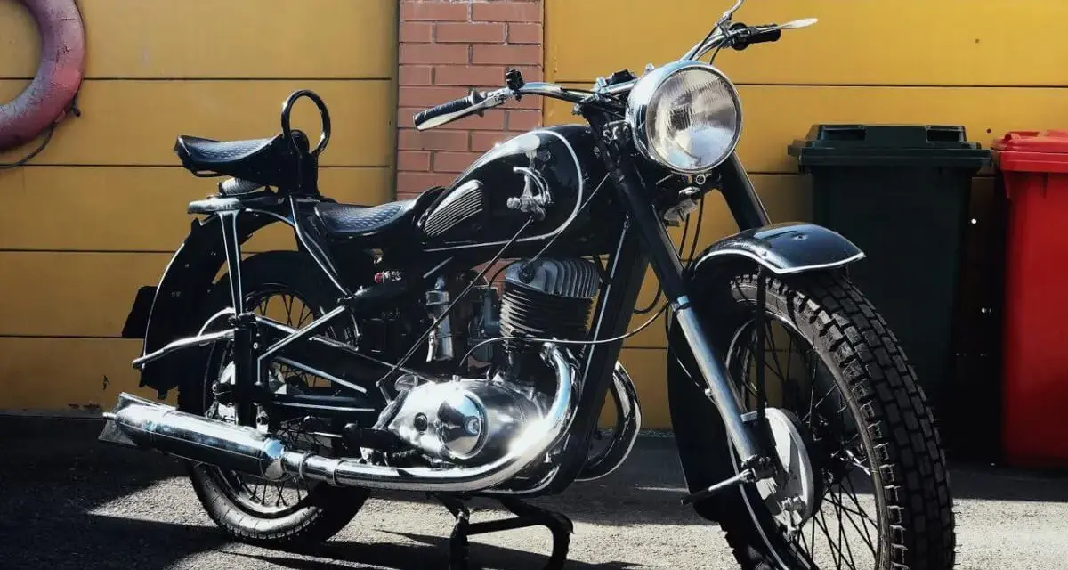 Klasyczny czarny motocykl, model iż 49, lśni w słońcu. Obok żółta brama i ceglana ściana.