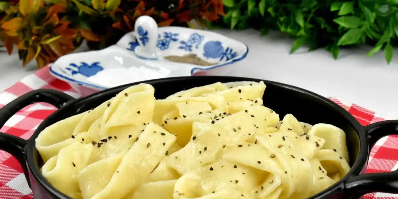 Pappardelle w kremowym sosie alfredo, posypane świeżo mielonym pieprzem, podane w czarnym naczyniu na czerwono-białej kratce.