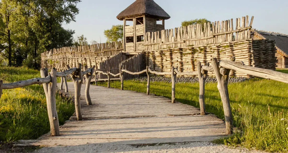 Drewniana kładka prowadzi do grodziska, jednego z historycznych miejsc w Polsce. Widać palisadę i wieżę obronną.