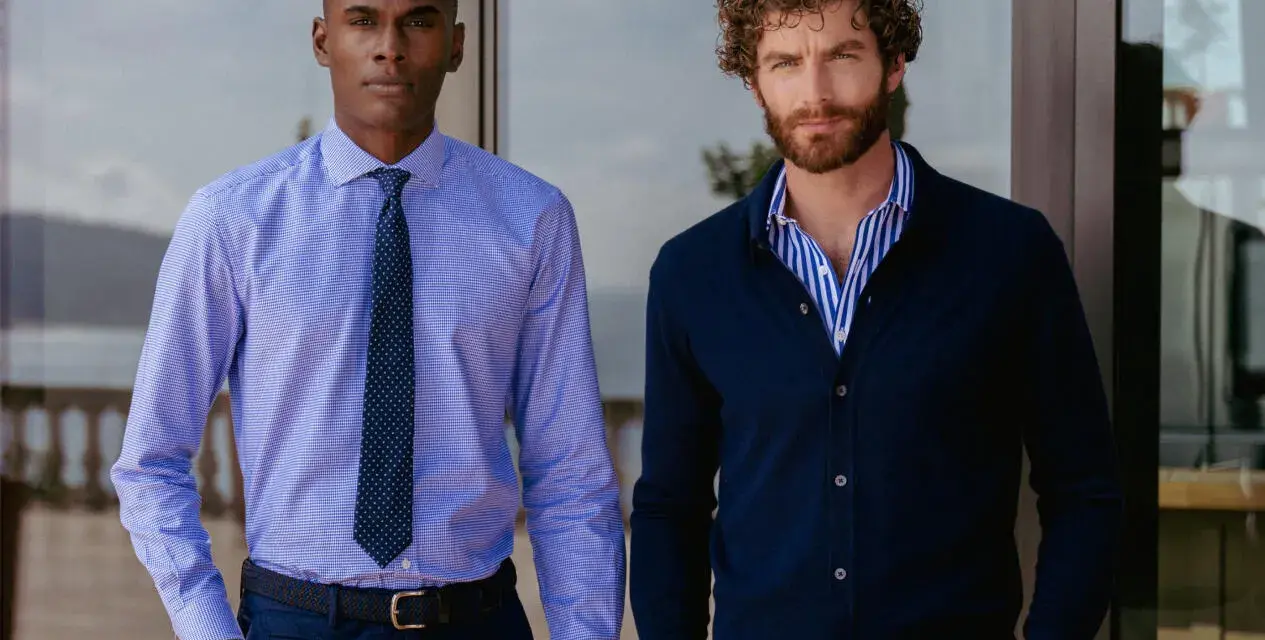 Dos hombres posan. Uno lleva una camisa de vestir azul con corbata, la camisa por dentro. El otro, una camisa de rayas azules y blancas con cárdigan azul oscuro, la camisa por fuera.
