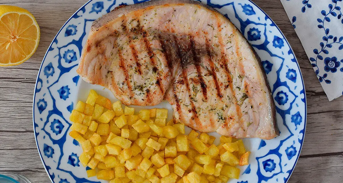 Delicioso emperador a la plancha con patatas fritas crujientes, servido en un plato con diseño azul y blanco. Un toque de limón completa esta apetitosa presentación.