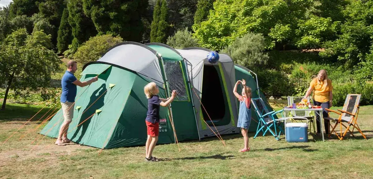 Radosny camping rodzinny: tata rozkłada namiot, dzieci bawią się piłką, mama przygotowuje posiłek.
