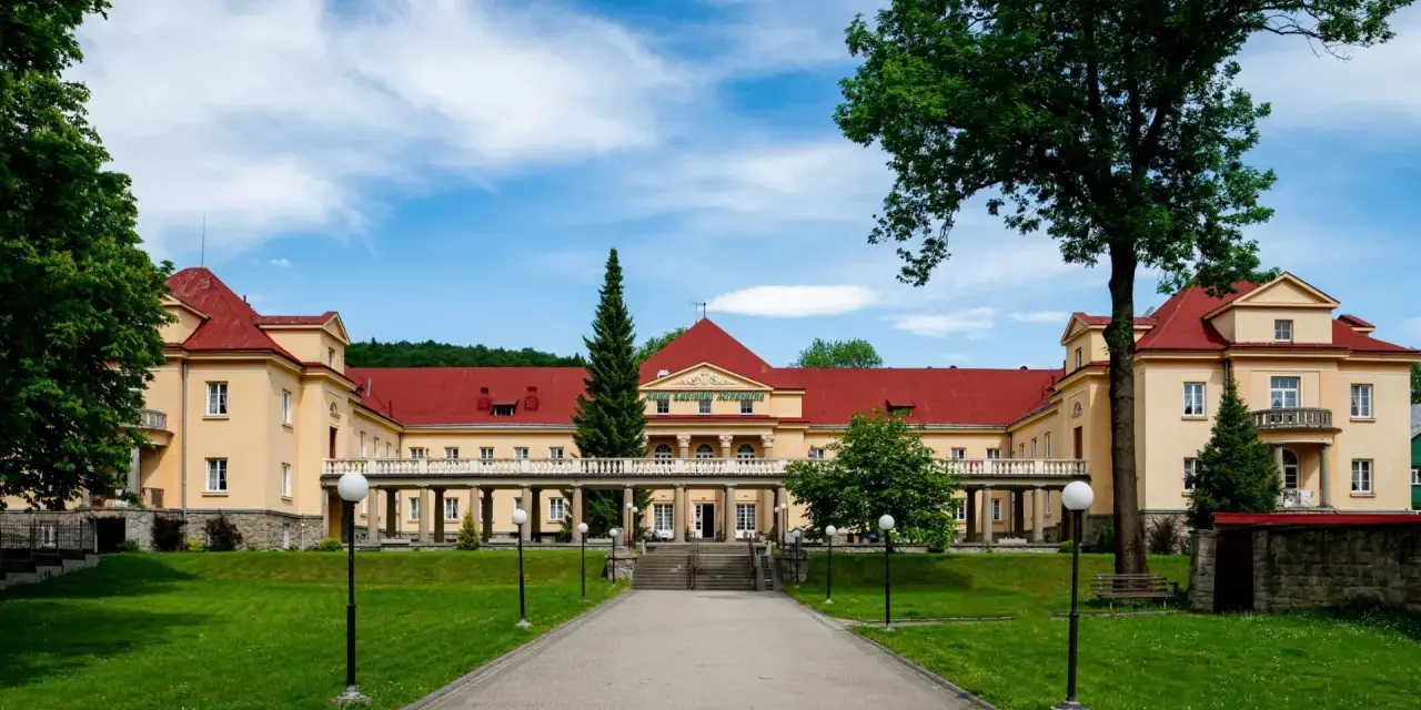Eleganckie, żółte budynki z czerwonymi dachami, przypominające stare łazienki Krynica Zdrój, otoczone zielonym ogrodem.