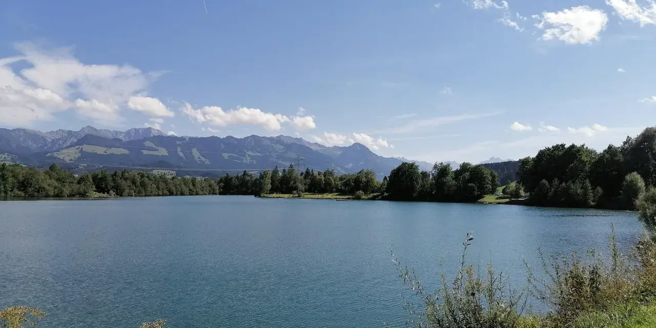 Blauer Himmel über dem Sonthofener See, umgeben von grünen Bäumen und majestätischen Bergen.