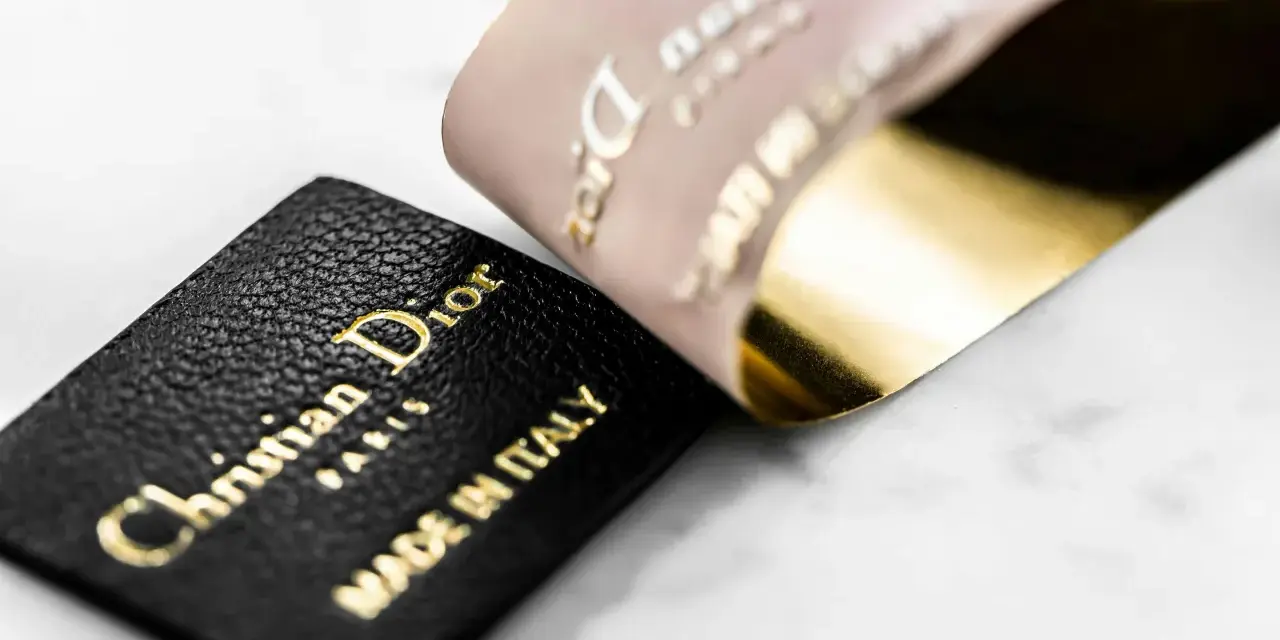 Etichetta in pelle nera con logo Christian Dior Paris e "Made in Italy" dorato, accanto a un nastro Dior francese con finitura oro lucido.