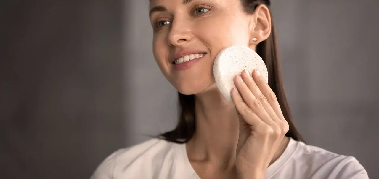 Une femme souriante utilise une éponge naturelle pour se laver le visage. Une méthode douce pour un nettoyage en douceur.