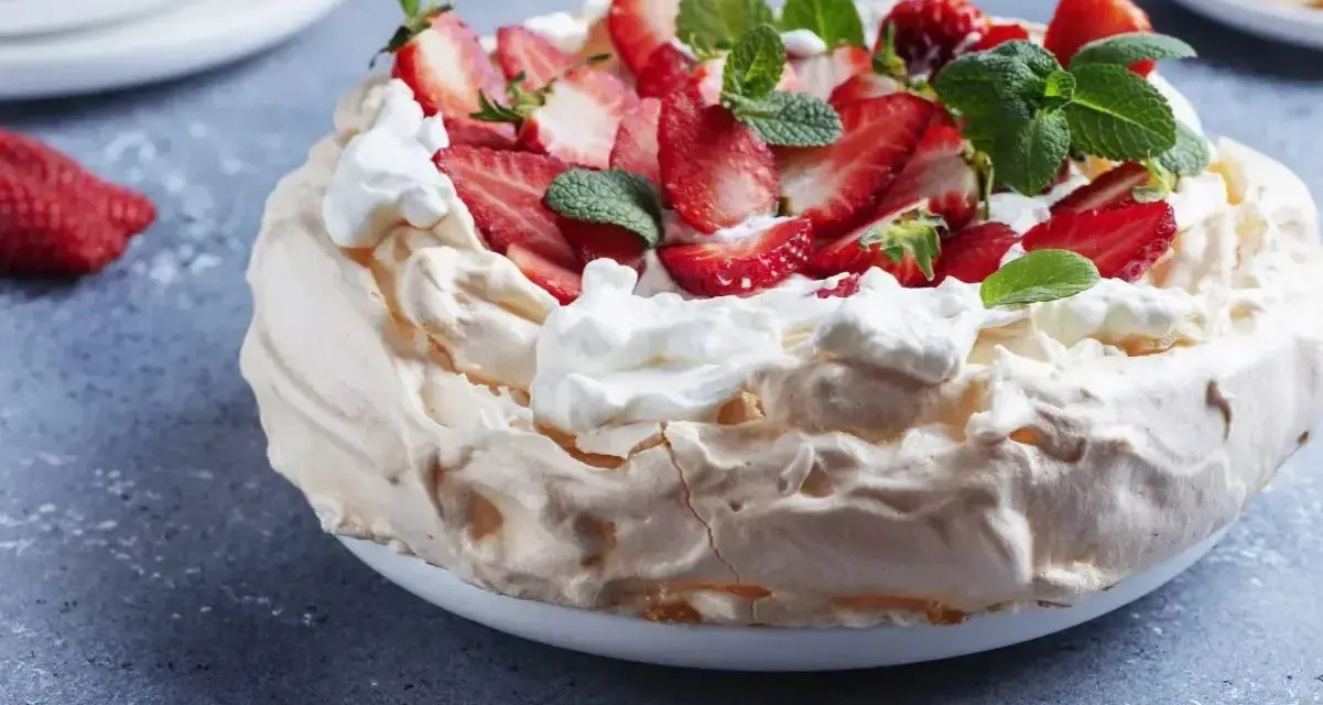 Pyszny tort bezowy z mascarpone i bitą śmietaną, ozdobiony świeżymi truskawkami i miętą.