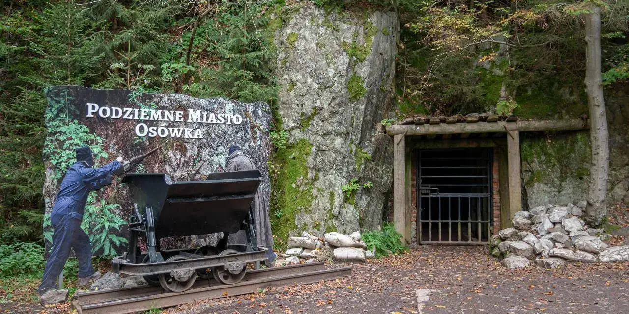 Wejście do Podziemnego Miasta Osówka, z wagonikiem i tablicą informacyjną. Widać skalne ściany i wejście do tunelu.