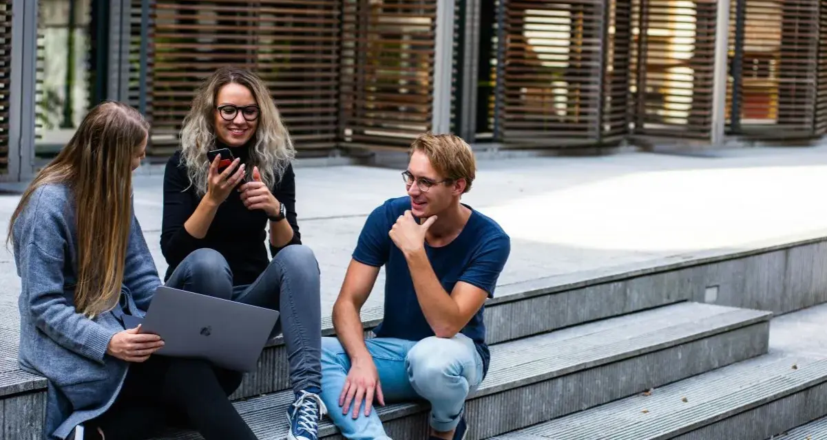 Trójka studentów, w tym jedna osoba na studiach lingwistycznych, siedzi na schodach, korzystając z laptopa i telefonu.