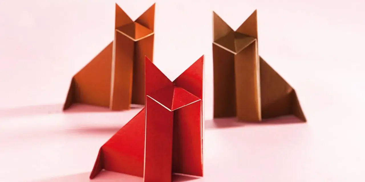 Drei origami Füchse, einer rot, zwei braun, sitzen auf rosa Untergrund.