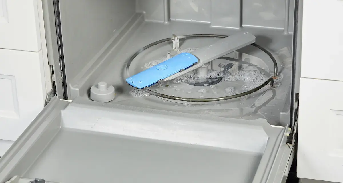 Water and suds fill the bottom of a dishwasher, with a blue spray arm component visible. This image shows a step in how to fix a clogged dishwasher.
