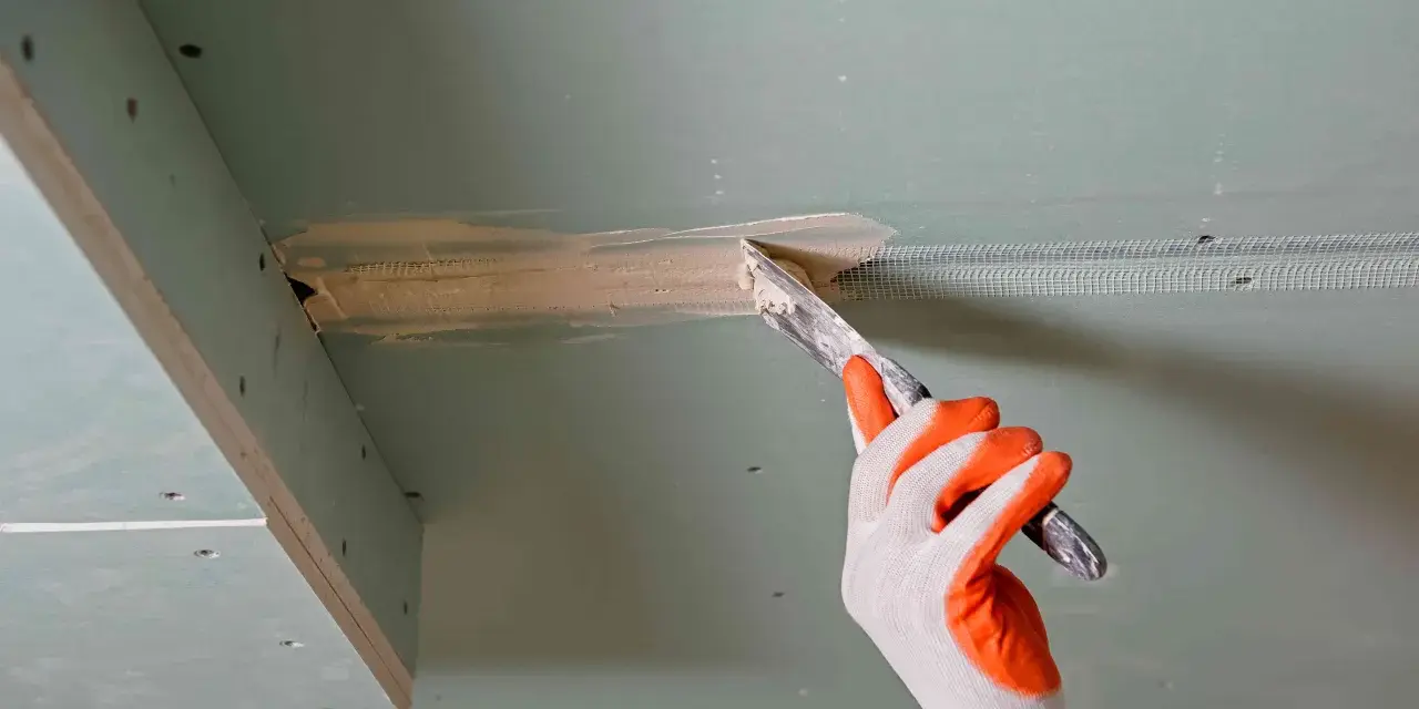Ręka w rękawicy nakłada masę szpachlową na połączenie płyt gipsowo-kartonowych, przygotowując je do dalszych prac. Klejenie płyt GK na ściany wymaga precyzji.